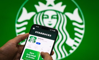 A person holds a smartphone displaying the Starbucks app with a large logo in the background.