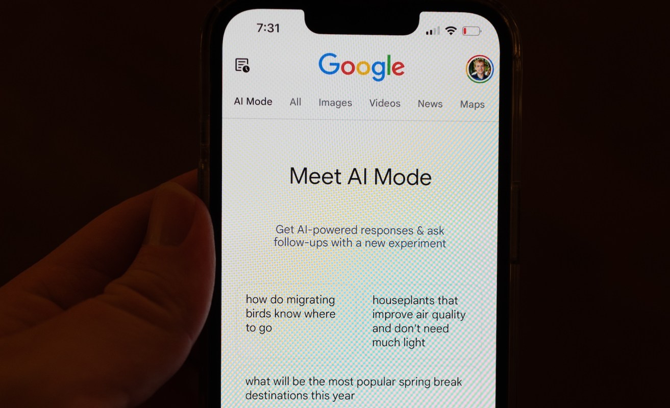 A smartphone displaying the Google search page with "Meet AI Mode" and related questions on the screen.