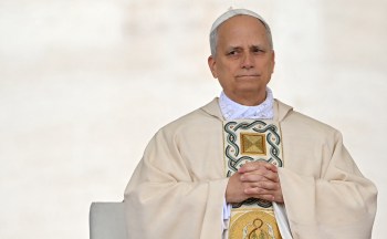 Pope Leo XIV in religious attire sits with hands clasped, looking thoughtfully, against a soft, neutral background.