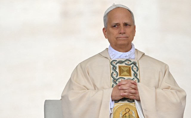 Pope Leo XIV in religious attire sits with hands clasped, looking thoughtfully, against a soft, neutral background.