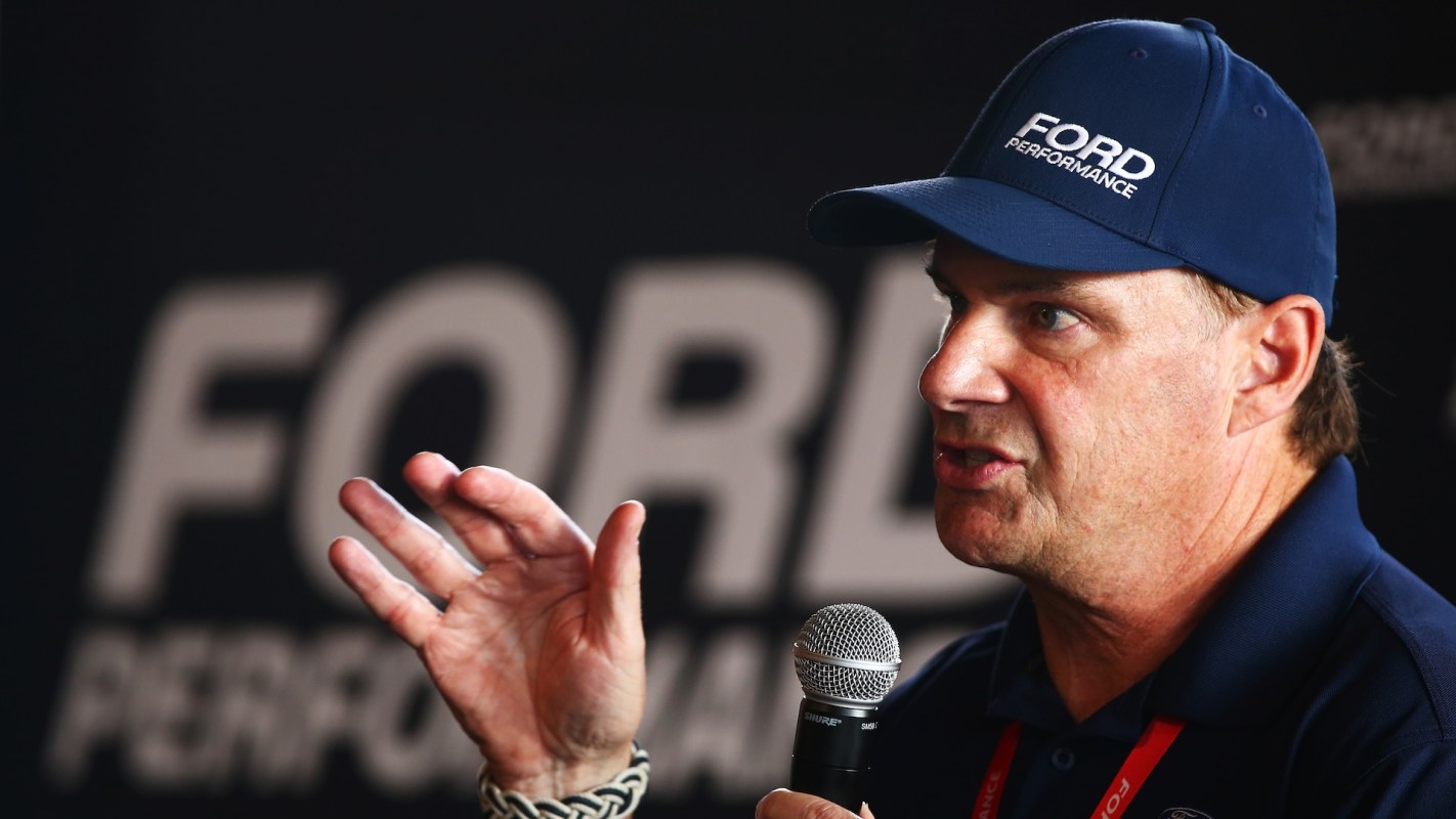 Ford CEO Jim Farley in a Ford cap speaks into a microphone, gesturing against a blurred Ford Performance background.