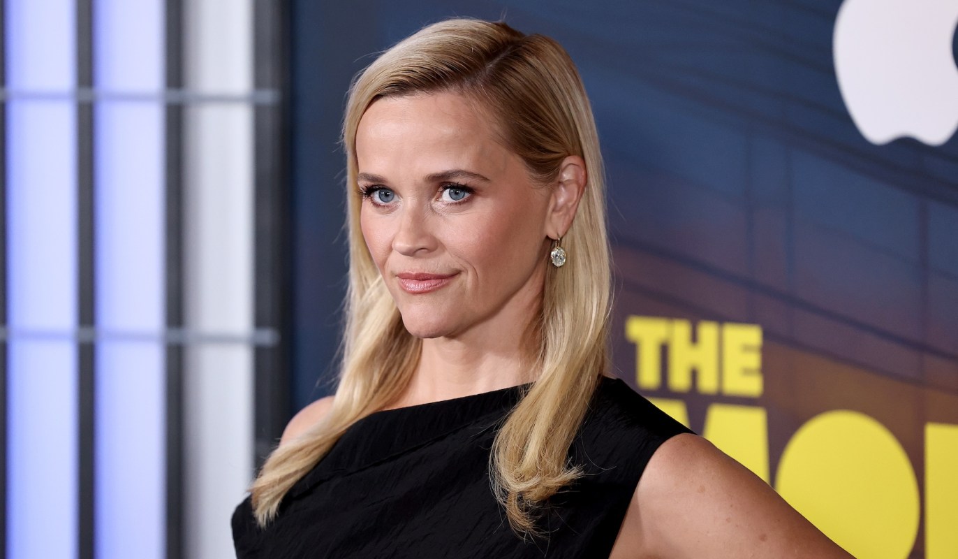Actress Reese Witherspoon, with long blonde hair wearing a black dress, poses confidently at a promotional event backdrop.