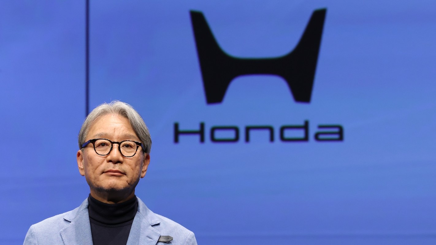 Toshihiro Mibe wears a light blue blazer and stands in front of a large Honda logo on a blue background.