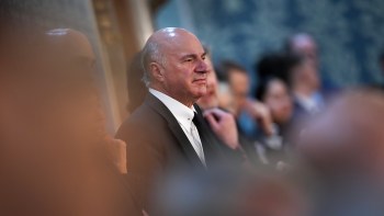 Kevin O'Leary in a black suit stands attentively in a blurred group setting.