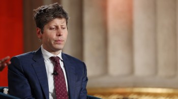 Sam Altman in a suit looks pensive while seated on stage during an event.