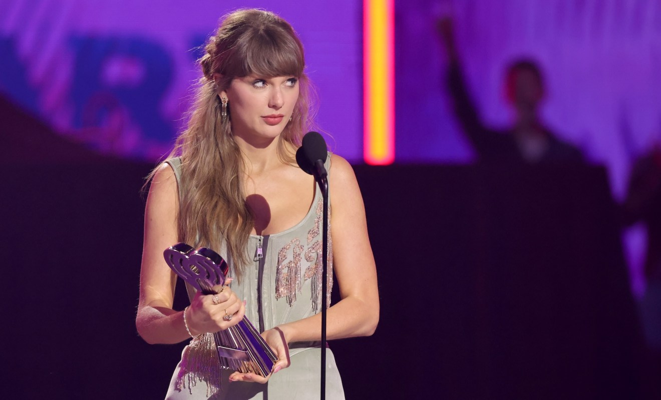 Taylor Swift stands at a microphone holding a trophy, looking thoughtful amid a colorful stage backdrop.