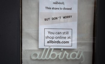 A closed Allbirds store window display featuring signs about online shopping availability.