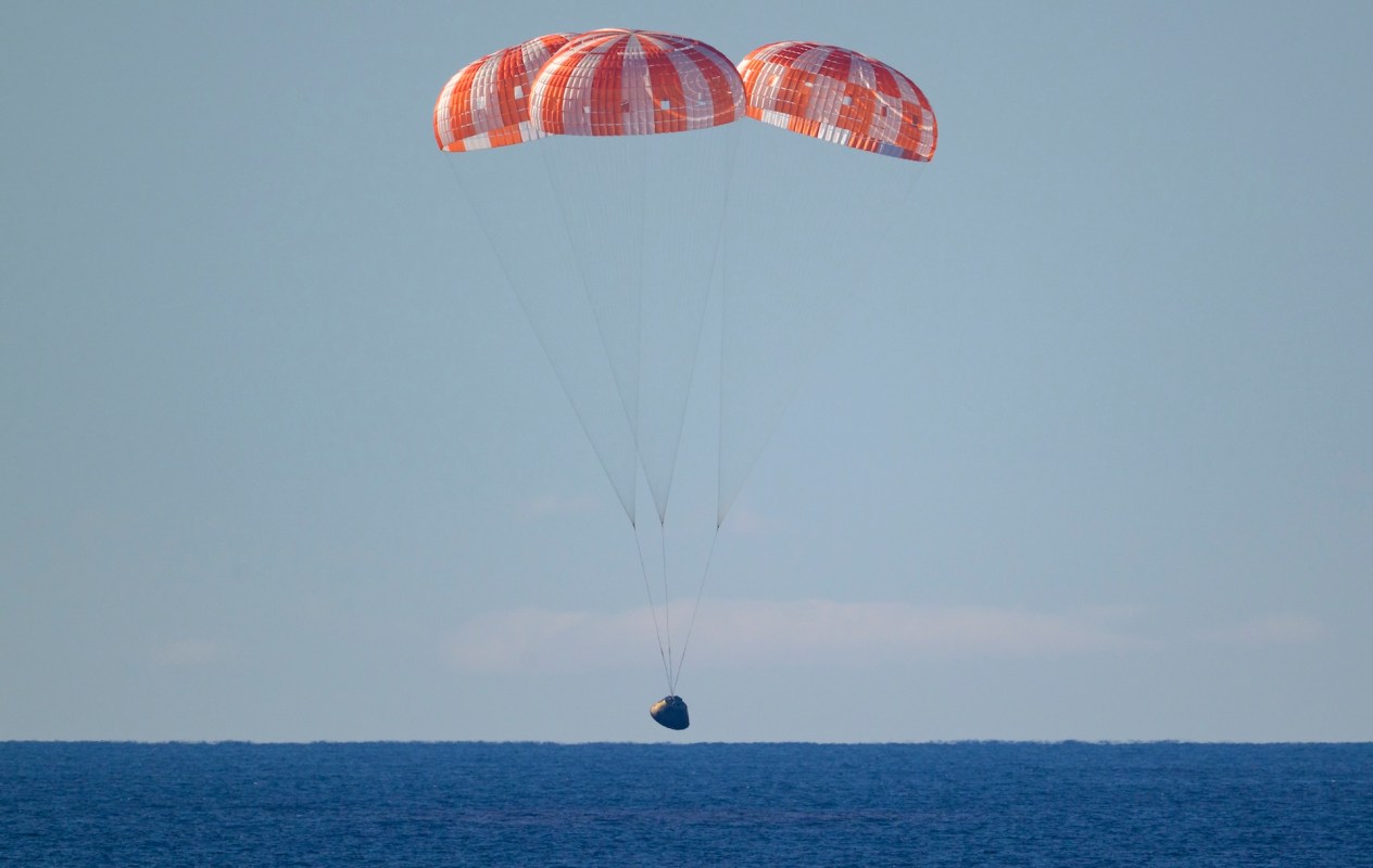 The Artemis 2 lunar capsule descends with three orange-and-white parachutes above a calm ocean.