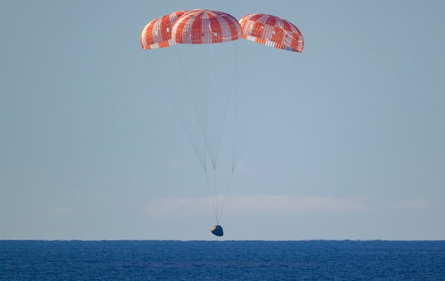 The Artemis 2 lunar capsule descends with three orange-and-white parachutes.