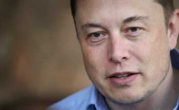 A close-up portrait of Elon Musk, wearing a blue collared shirt and with slight stubble.
