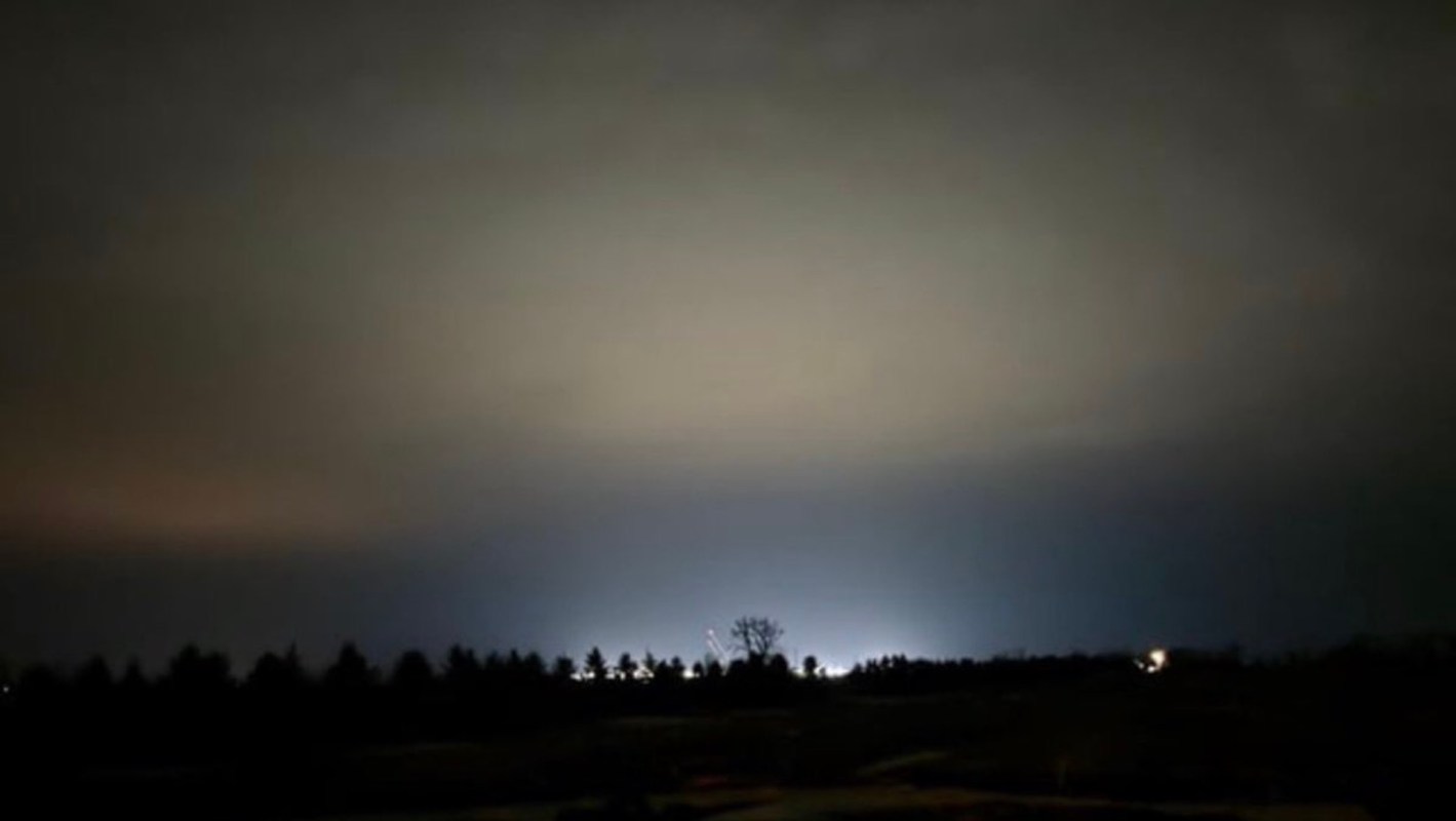 A dimly lit sky with subtle color gradients and distant lights peeking through silhouetted trees from a data center.