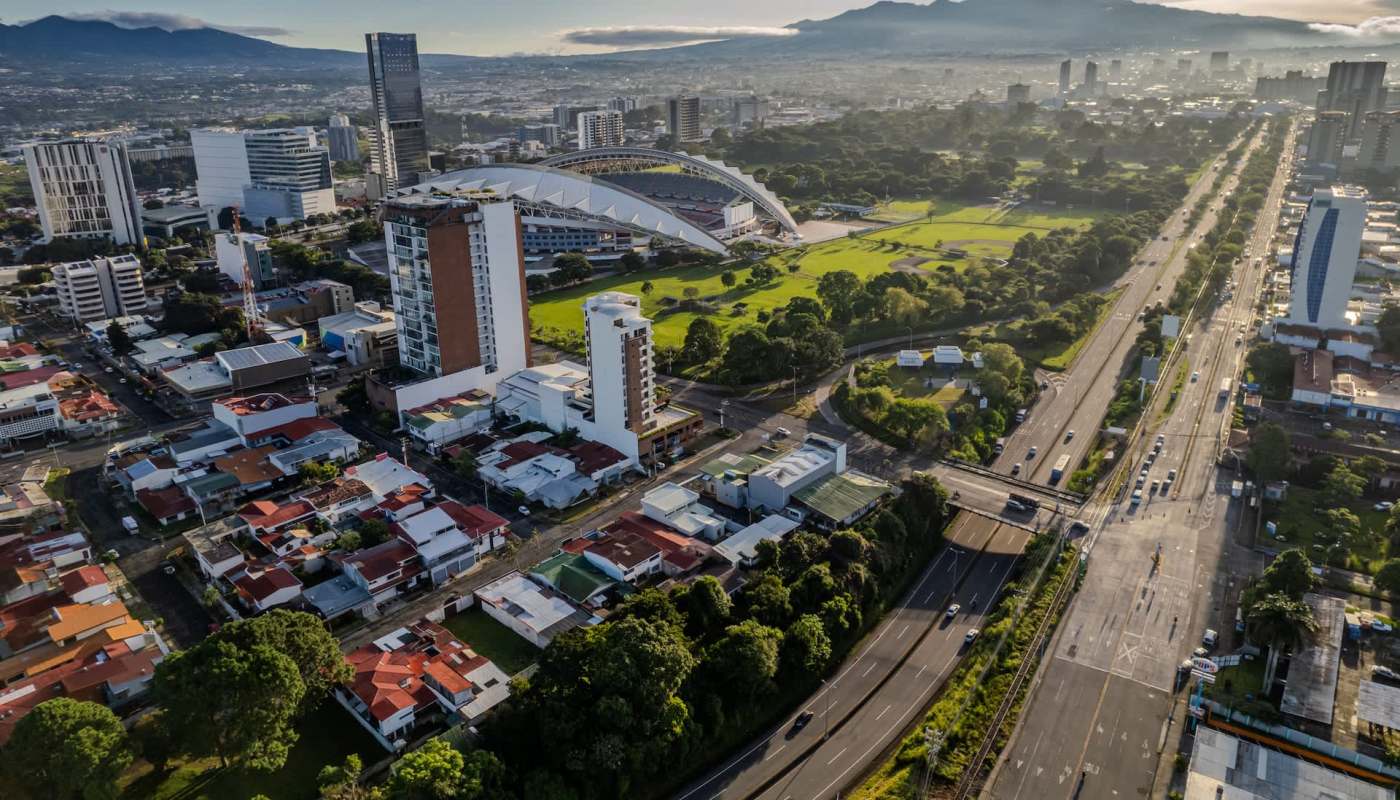 Aerial view of a city with modern buildings, a stadium, green spaces, and highways lined with traffic.