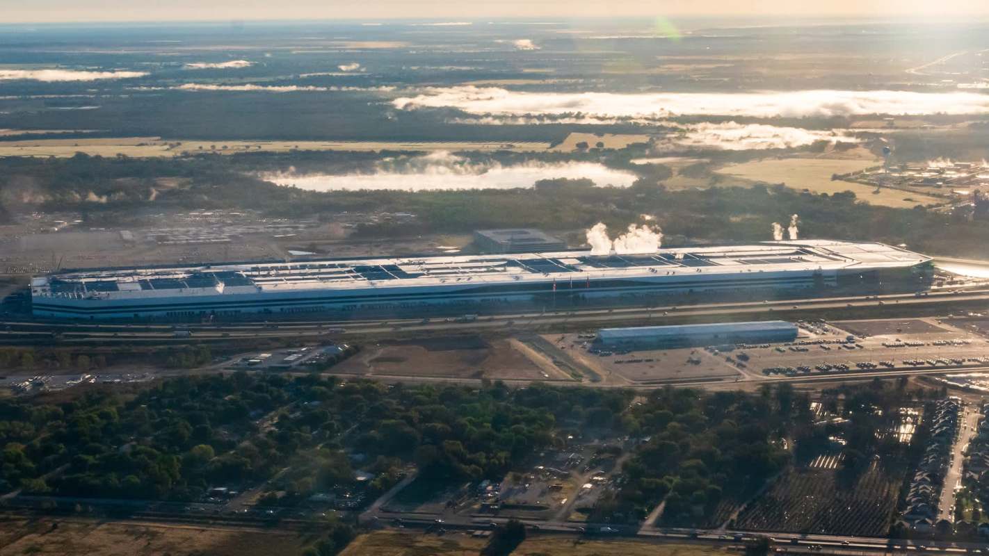 Aerial view of a the Tesla Gigafactory in Austin, Texas, with smoke rising, surrounded by greenery and parking lots.