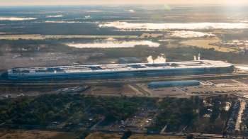 Aerial view of a the Tesla Gigafactory in Austin, Texas, with smoke rising, surrounded by greenery and parking lots.