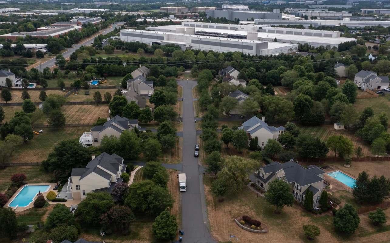 An aerial view of a suburban neighborhood featuring houses, trees, and swimming pools, with a data center in the background.