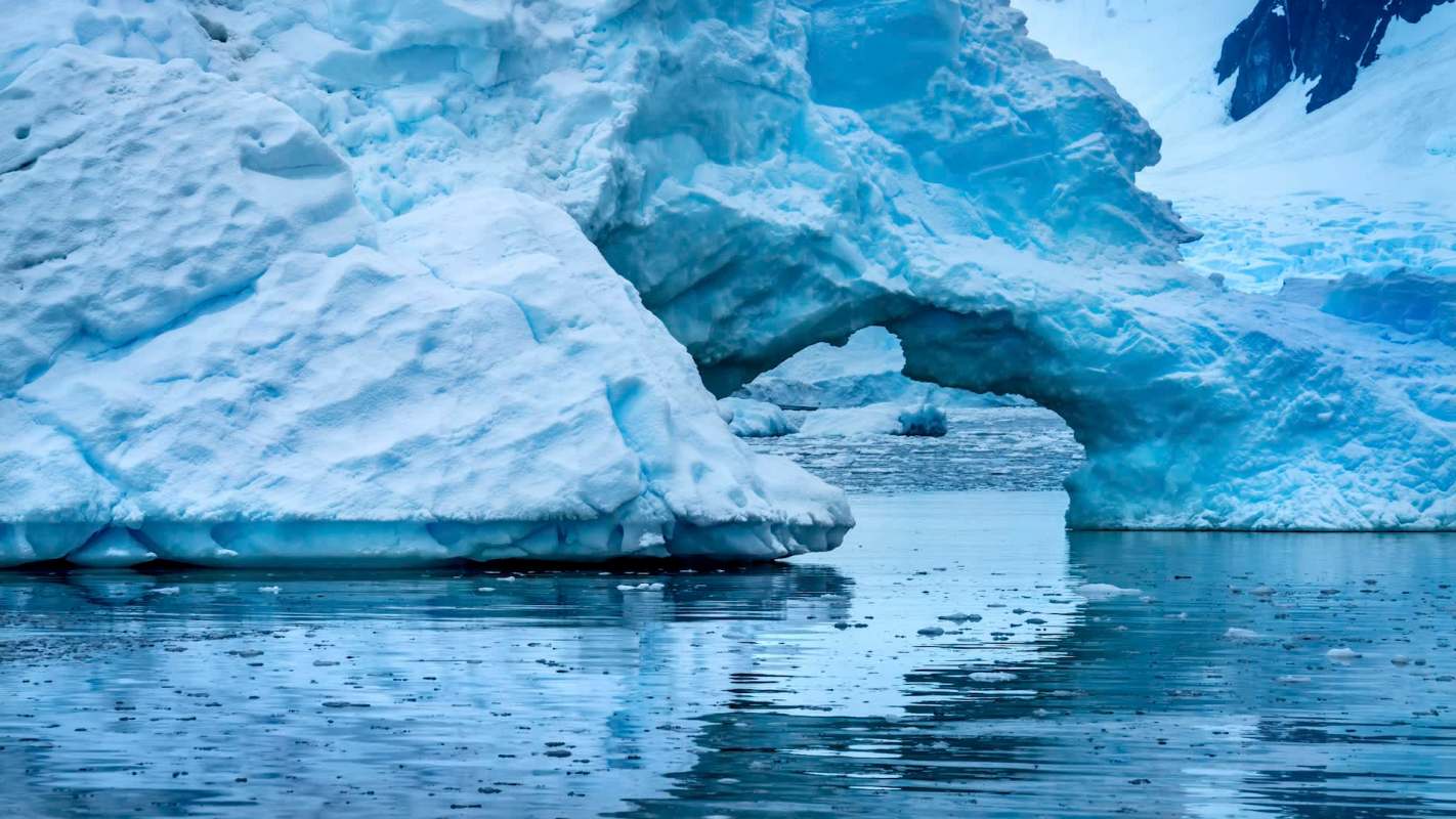 A large icy arch in Antarctica is surrounded by floating icebergs in a calm, reflective water body.