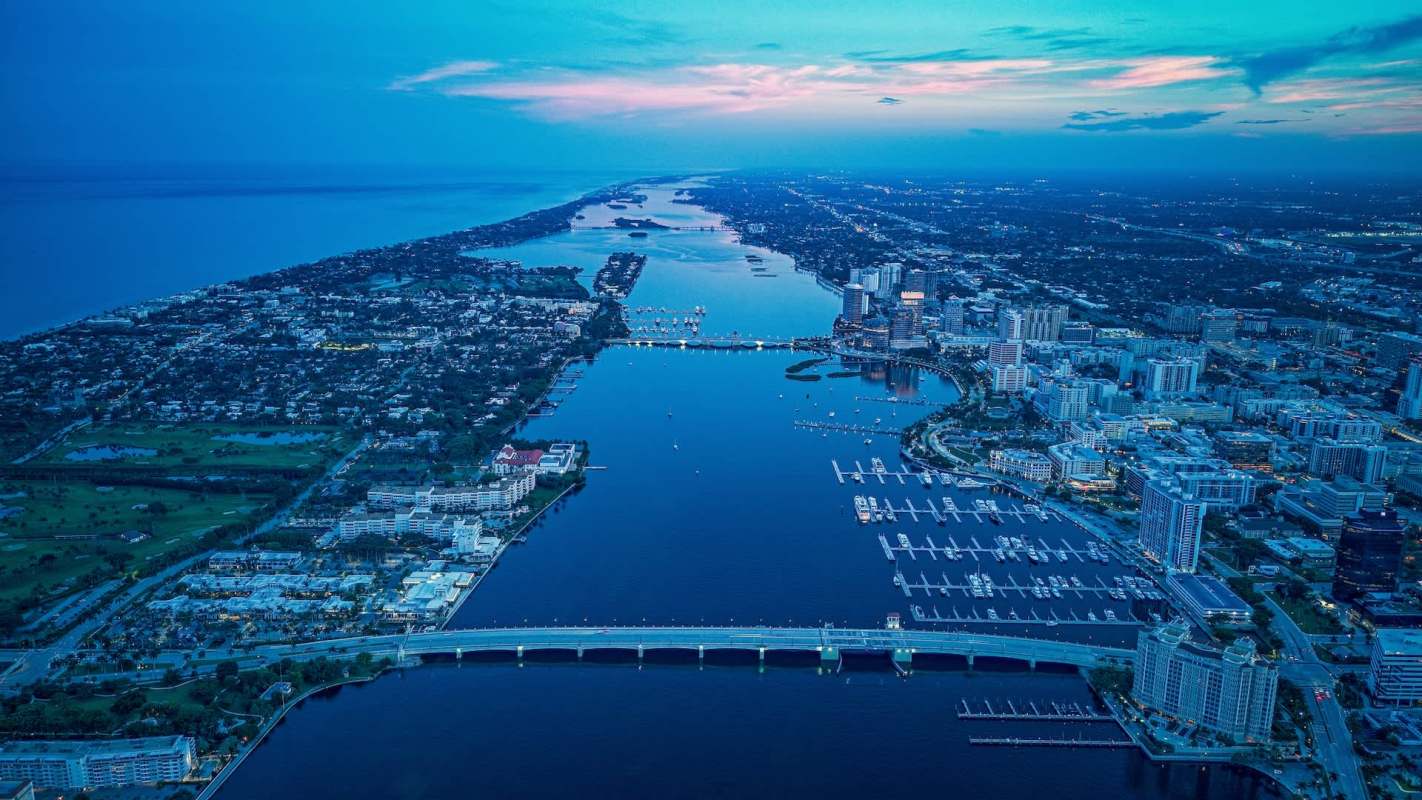 Aerial view of a waterfront city at twilight, showcasing buildings, bridges, and marina boats.