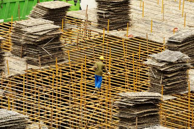 A worker navigates a scaffolding structure on a construction site surrounded by stacks of wooden materials.