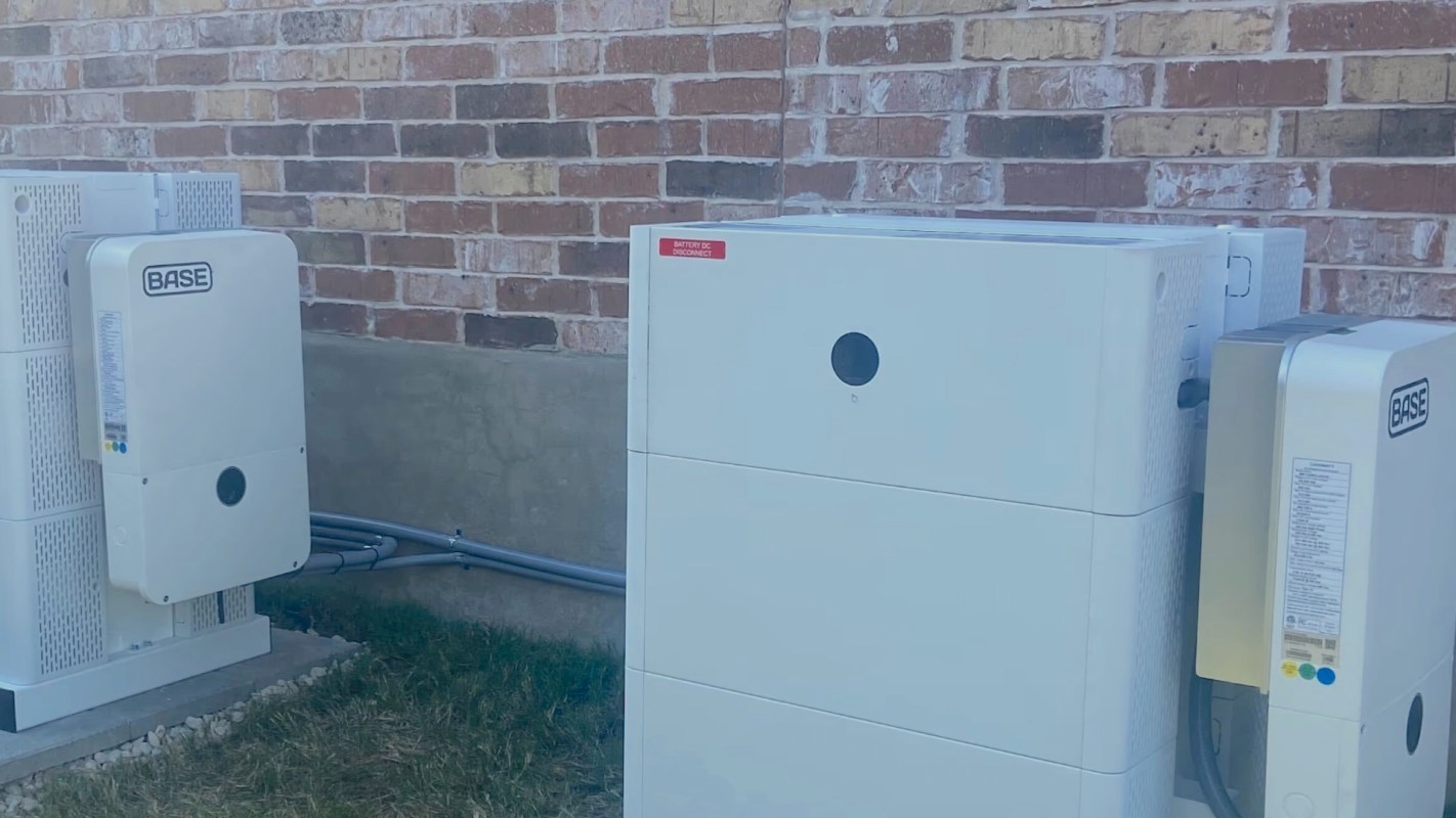 A white battery storage unit sits against a brick wall with visible electrical connections and grass at its base.