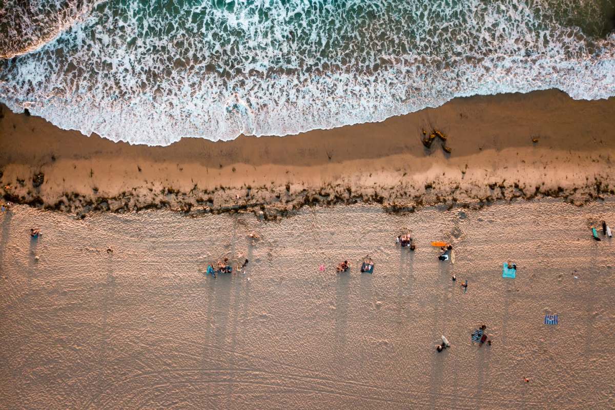 An aerial view of a sandy beach with people relaxing near the shoreline and gentle waves lapping at the sand.