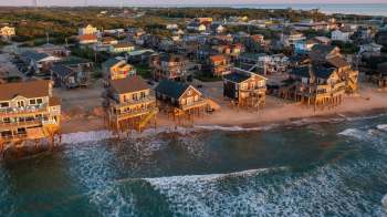 Aerial view of beachfront houses on stilts, with waves lapping at the shore and greenery in the background.