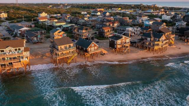 Aerial view of beachfront houses on stilts, with waves lapping at the shore and greenery in the background.