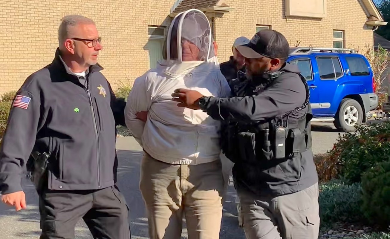 Two police officers escort a handcuffed person wearing a beekeeping suit outside a house.