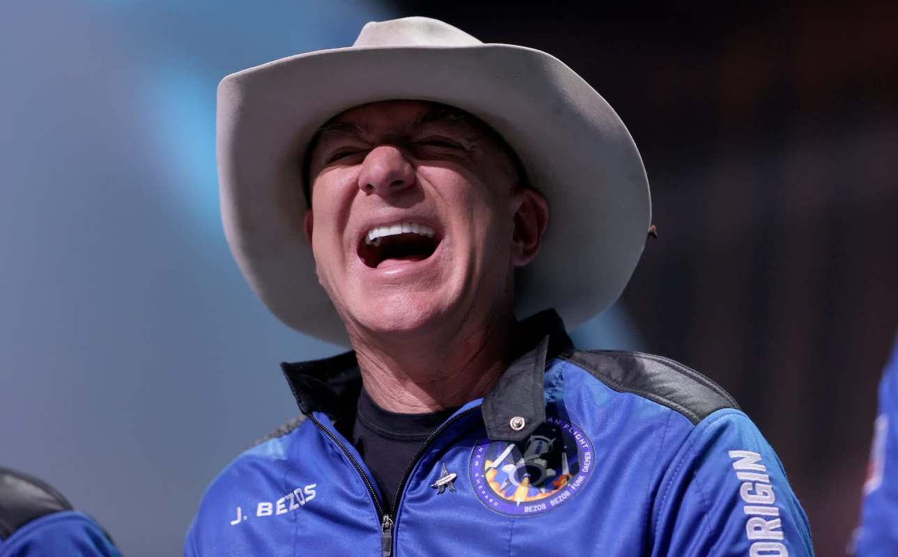 Jeff Bezos in a cowboy hat joyfully laughing while wearing a blue jacket with a Blue Origin logo.