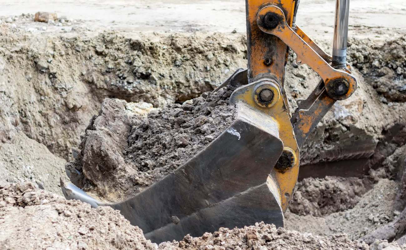 A close-up of a construction excavator bucket lifting dirt from a large trench.