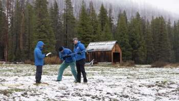 Three researchers in blue jackets are examining snowpack data near a wooden cabin.