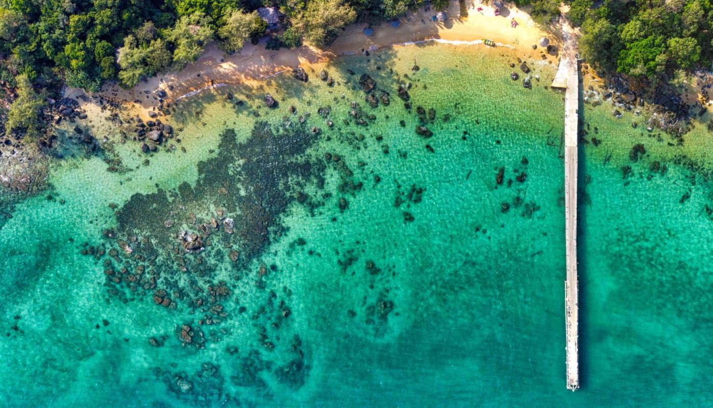 An aerial view of a sandy beach with a wooden pier extending into clear turquoise water.