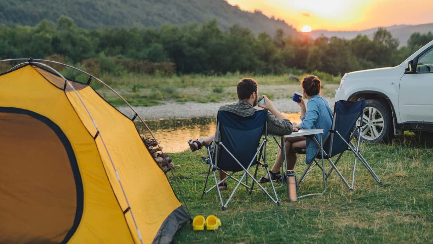 A couple relaxes by a river at sunset, seated in camping chairs next to a yellow tent and a white SUV.
