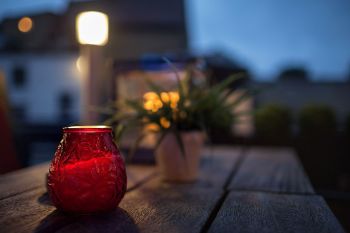 A red glass candle holder on a wooden table with a plant and soft evening lighting in the background.