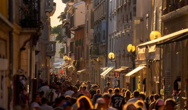A bustling narrow street filled with evening crowds, warm sunlight illuminating historic facades, shop signs, and lively summer atmosphere.