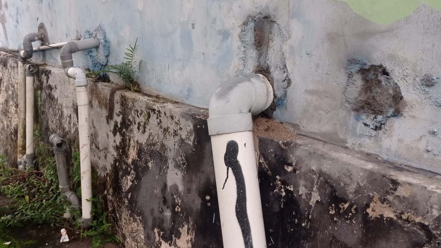 A weathered wall features multiple pipes, some with moss, and a graffiti-like figure on a white pipe.