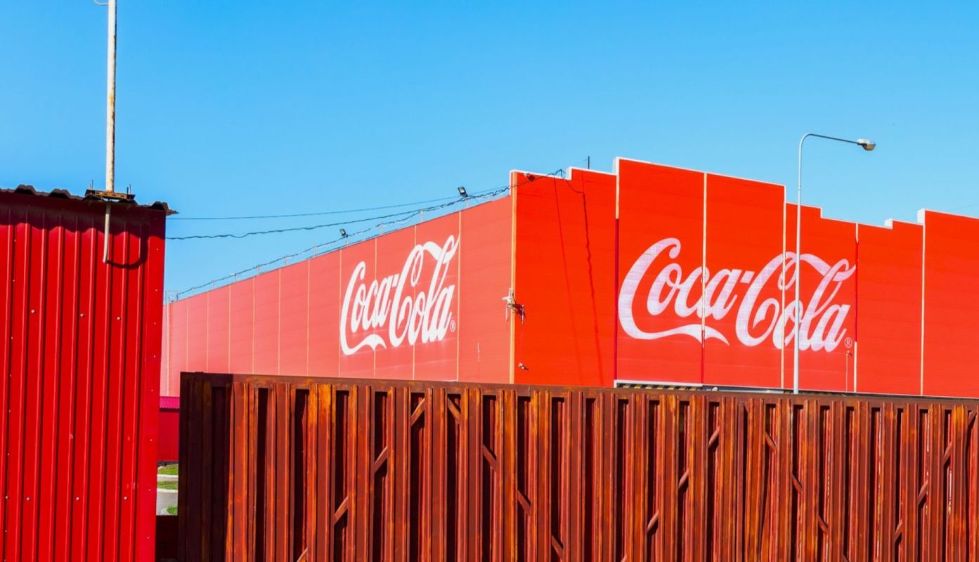 A bright red Coca-Cola advertisement on a building, partially obscured by a wooden fence.