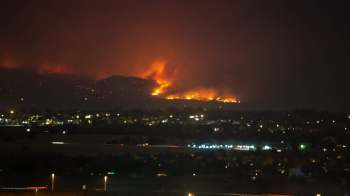 A nighttime view of a wildfire burning on a hillside, with glowing flames and smoke against a dark sky.