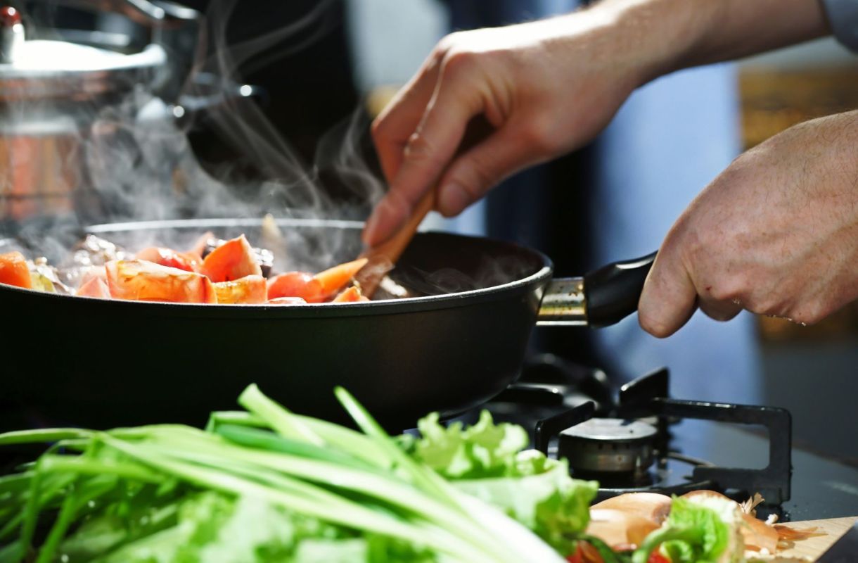 A person stirring chopped vegetables in a steaming frying pan, with fresh greens in the foreground.