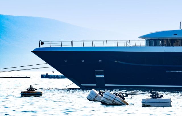 A luxury yacht anchored near floating buoys on calm waters, with mountains in the background.