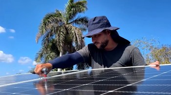 A man wearing a hat works on a solar panel under a clear blue sky with palm trees in the background.