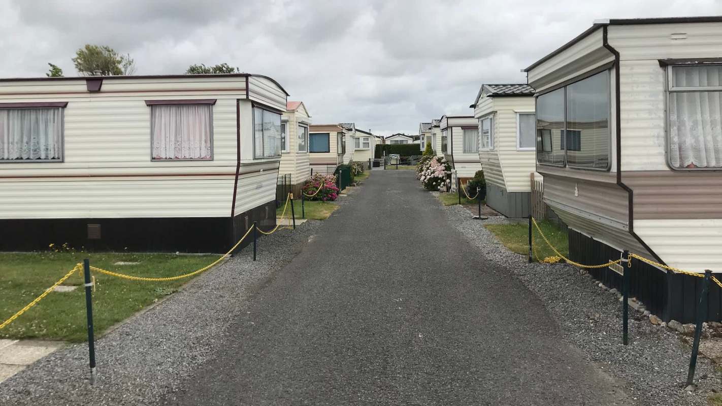 A narrow gravel pathway in a mobile home community lined with several static caravans and flower gardens under a cloudy sky.
