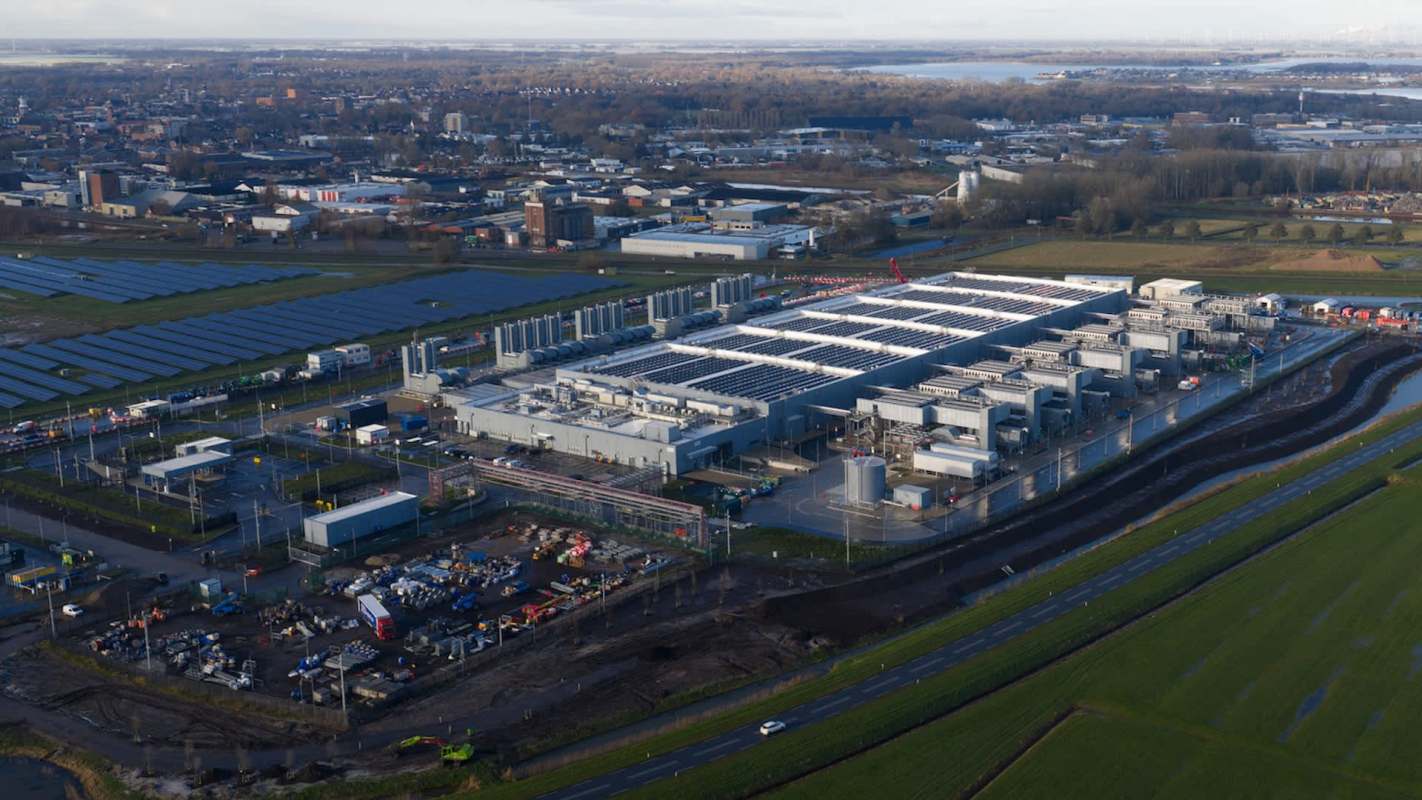 An aerial view of a large data center surrounded by solar panels, a rural landscape, and buildings.