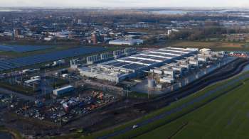 An aerial view of a large data center surrounded by solar panels, a rural landscape, and buildings.