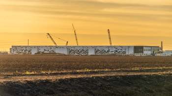 A large industrial building under construction with cranes against a golden sunset sky.