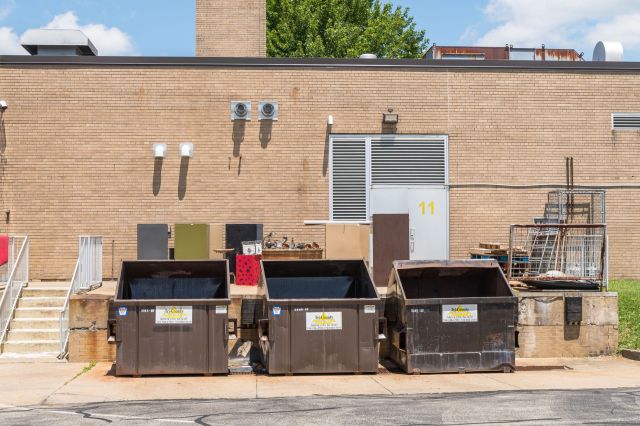 A row of three dumpsters stands against a brick wall with scattered furniture and debris nearby.