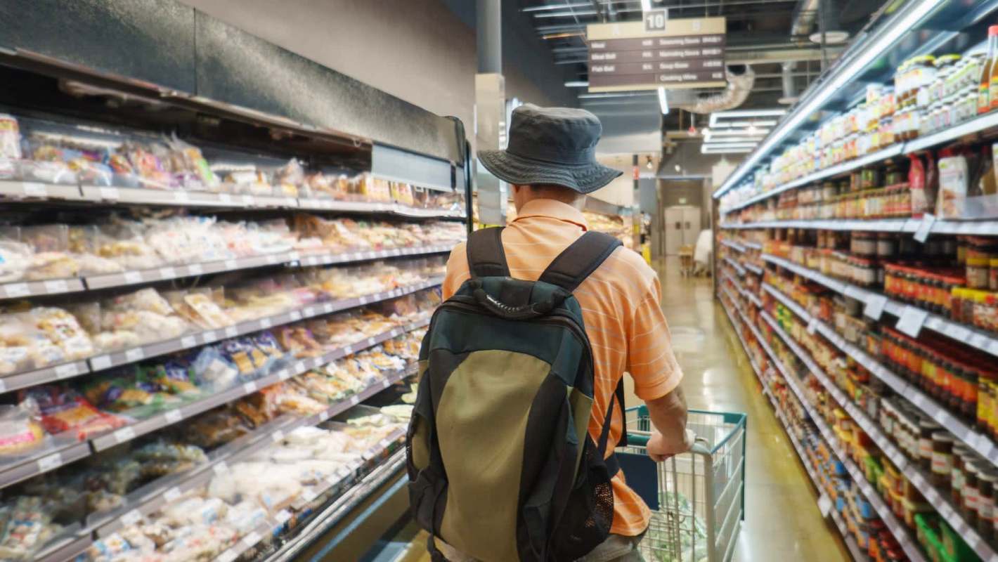 A person wearing a bucket hat pushes a shopping cart down an aisle filled with various packaged foods.