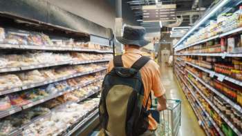 A person wearing a bucket hat pushes a shopping cart down an aisle filled with various packaged foods.