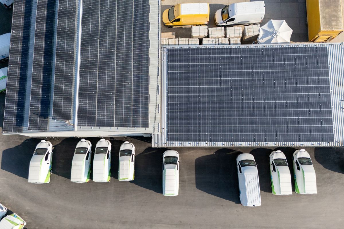 An aerial view of a parking area with white delivery van EVs and solar panel-covered roofs.