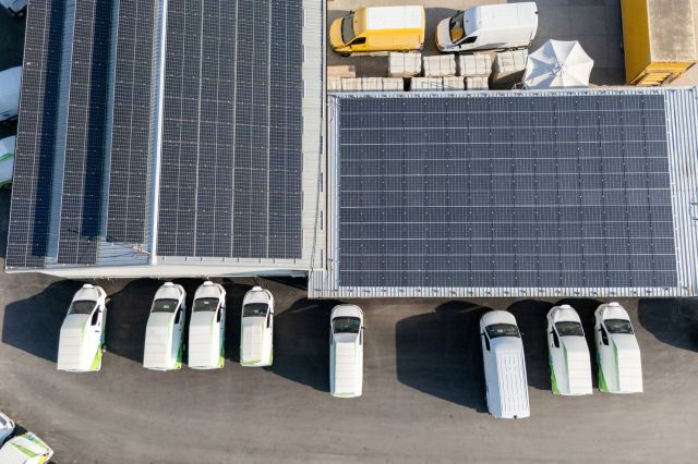 An aerial view of a parking area with white delivery van EVs and solar panel-covered roofs.