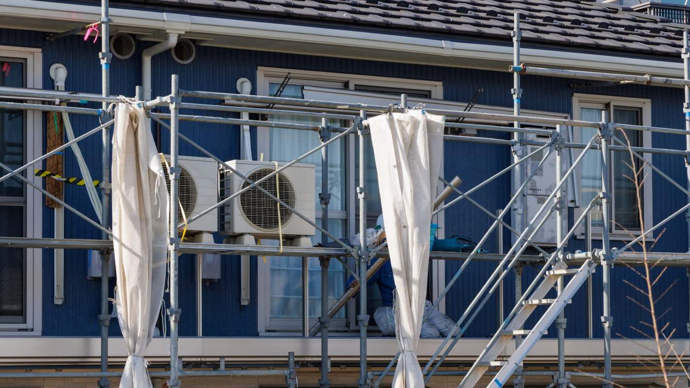A worker in protective gear is painting the exterior of a building using scaffolding and air conditioning units are visible.